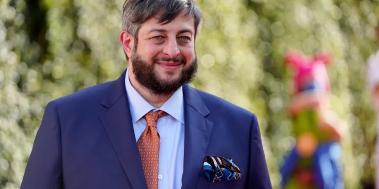 Eugene Mirman, wearing a blue suit, orange tie, and pocket square, smiles at the camera.
