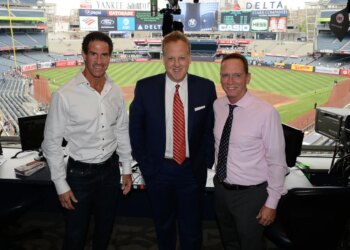 Three men posing for a photo in a broadcast booth overlooking Yankee Stadium.