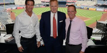 Three men posing for a photo in a broadcast booth overlooking Yankee Stadium.