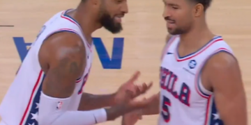 Two basketball players in white jerseys with red and blue accents, facing each other on the court.