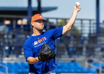 A baseball player in a blue Mets shirt and orange hat throwing a baseball during spring training.