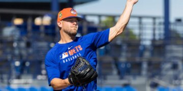 A baseball player in a blue Mets shirt and orange hat throwing a baseball during spring training.