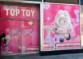 Storefront of the new Top Toy store in Times Square, with pink signs reading "TOP TOY" and "Coming Soon TRENDY TOY" on the left doors, and an illustration of a large cartoon doll in a fluffy pink outfit on the right window.