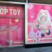 Storefront of the new Top Toy store in Times Square, with pink signs reading "TOP TOY" and "Coming Soon TRENDY TOY" on the left doors, and an illustration of a large cartoon doll in a fluffy pink outfit on the right window.