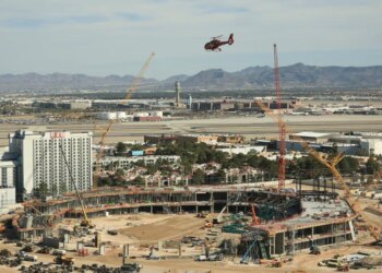 Construction site of a new ballpark in Las Vegas, with a helicopter flying over, a hotel in the foreground, and an airport with mountains in the background.