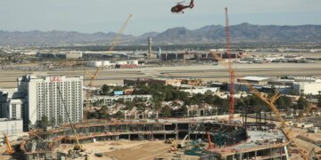Construction site of a new ballpark in Las Vegas, with a helicopter flying over, a hotel in the foreground, and an airport with mountains in the background.