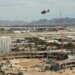 Construction site of a new ballpark in Las Vegas, with a helicopter flying over, a hotel in the foreground, and an airport with mountains in the background.