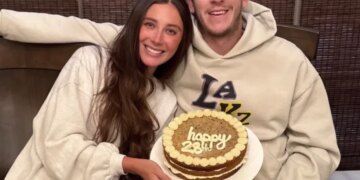 A couple smiles while holding a "happy 23rd" birthday cake.