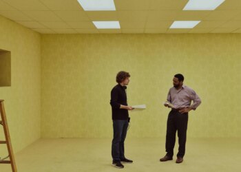 A image of two men on a film set made to look like the interior of a bland, beige office building.