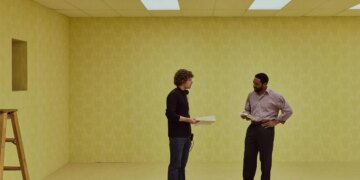 A image of two men on a film set made to look like the interior of a bland, beige office building.