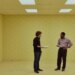 A image of two men on a film set made to look like the interior of a bland, beige office building.