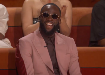 Draymond Green laughs while wearing dark round sunglasses and a pink suit over a dark shirt and tie, sitting in a red chair.