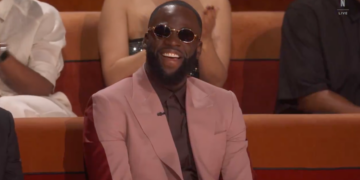 Draymond Green laughs while wearing dark round sunglasses and a pink suit over a dark shirt and tie, sitting in a red chair.
