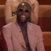 Draymond Green laughs while wearing dark round sunglasses and a pink suit over a dark shirt and tie, sitting in a red chair.