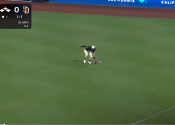 A baseball player in a white and dark blue uniform with a red glove fields a baseball on a green field during a game with a scoreboard showing the Cardinals vs. Padres, 5th inning, 0-0 score.