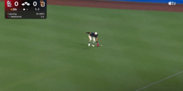 A baseball player in a white and dark blue uniform with a red glove fields a baseball on a green field during a game with a scoreboard showing the Cardinals vs. Padres, 5th inning, 0-0 score.