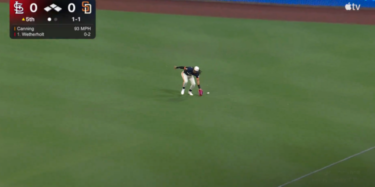 A baseball player in a white and dark blue uniform with a red glove fields a baseball on a green field during a game with a scoreboard showing the Cardinals vs. Padres, 5th inning, 0-0 score.