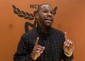Floyd Mayweather speaking and gesturing with both hands, wearing a black jacket with a repeating pattern, against an orange background with a black MCM logo.