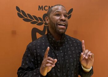 Floyd Mayweather speaking and gesturing with both hands, wearing a black jacket with a repeating pattern, against an orange background with a black MCM logo.