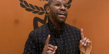 Floyd Mayweather speaking and gesturing with both hands, wearing a black jacket with a repeating pattern, against an orange background with a black MCM logo.