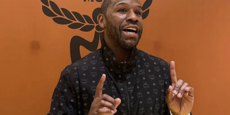 Floyd Mayweather speaking and gesturing with both hands, wearing a black jacket with a repeating pattern, against an orange background with a black MCM logo.