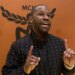 Floyd Mayweather speaking and gesturing with both hands, wearing a black jacket with a repeating pattern, against an orange background with a black MCM logo.