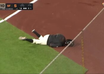 A baseball ball girl in a helmet has fallen onto the grass and dirt of the field.