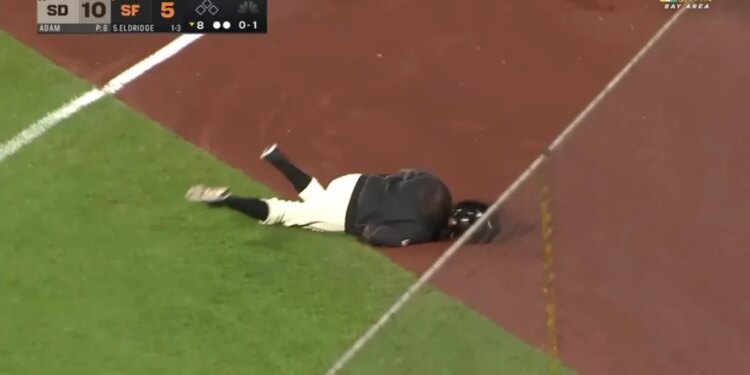 A baseball ball girl in a helmet has fallen onto the grass and dirt of the field.