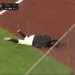 A baseball ball girl in a helmet has fallen onto the grass and dirt of the field.