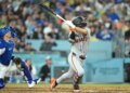 A San Francisco Giants batter in a grey uniform swinging a bat with a Los Angeles Dodgers catcher in blue gear watching from behind.