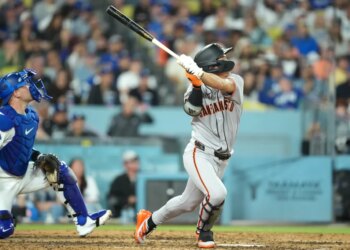 A San Francisco Giants batter in a grey uniform swinging a bat with a Los Angeles Dodgers catcher in blue gear watching from behind.