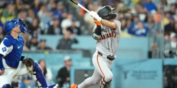 A San Francisco Giants batter in a grey uniform swinging a bat with a Los Angeles Dodgers catcher in blue gear watching from behind.