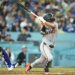 A San Francisco Giants batter in a grey uniform swinging a bat with a Los Angeles Dodgers catcher in blue gear watching from behind.