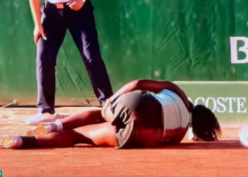 A tennis player in a black and white outfit is lying on the clay court, seemingly injured, while two officials stand over her.