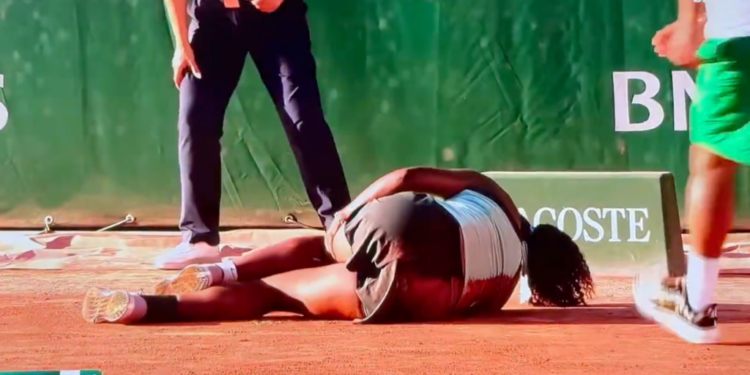 A tennis player in a black and white outfit is lying on the clay court, seemingly injured, while two officials stand over her.