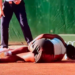 A tennis player in a black and white outfit is lying on the clay court, seemingly injured, while two officials stand over her.