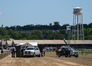 A horse receives medical attention inside a black tent on a dirt racetrack with a water tower in the background.