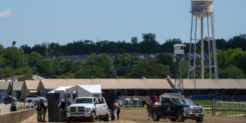 A horse receives medical attention inside a black tent on a dirt racetrack with a water tower in the background.