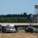 A horse receives medical attention inside a black tent on a dirt racetrack with a water tower in the background.