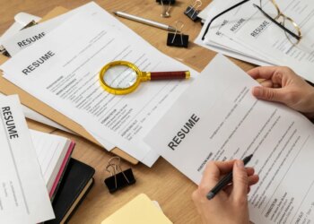 A person reviewing a resume, with other resumes, office supplies, and a magnifying glass on a wooden desk.