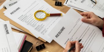 A person reviewing a resume, with other resumes, office supplies, and a magnifying glass on a wooden desk.