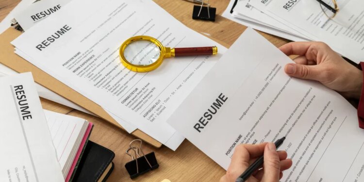 A person reviewing a resume, with other resumes, office supplies, and a magnifying glass on a wooden desk.