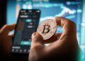 A person holds a Bitcoin coin while looking at a cryptocurrency trading app on a smartphone, with charts and graphs on a blurred screen in the background.