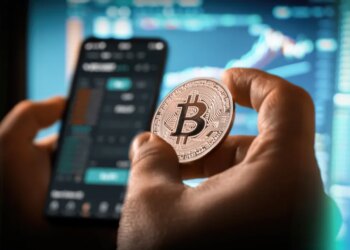 A person holds a Bitcoin coin while looking at a cryptocurrency trading app on a smartphone, with charts and graphs on a blurred screen in the background.
