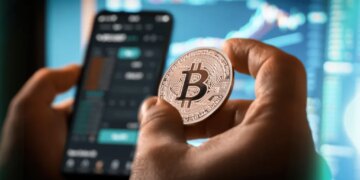 A person holds a Bitcoin coin while looking at a cryptocurrency trading app on a smartphone, with charts and graphs on a blurred screen in the background.