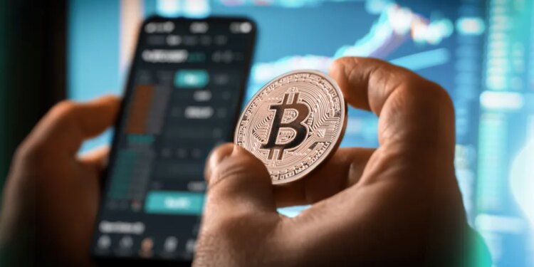 A person holds a Bitcoin coin while looking at a cryptocurrency trading app on a smartphone, with charts and graphs on a blurred screen in the background.