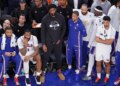 Philadelphia 76ers center Joel Embiid sits on the bench in plain clothes with teammates during a game.