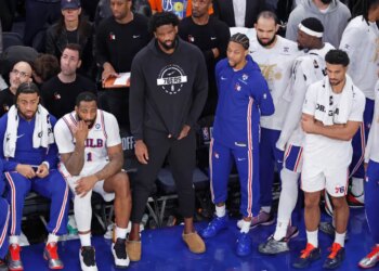 Philadelphia 76ers center Joel Embiid sits on the bench in plain clothes with teammates during a game.