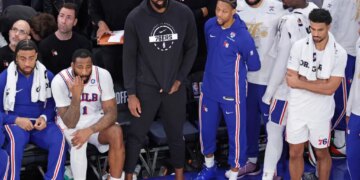 Philadelphia 76ers center Joel Embiid sits on the bench in plain clothes with teammates during a game.