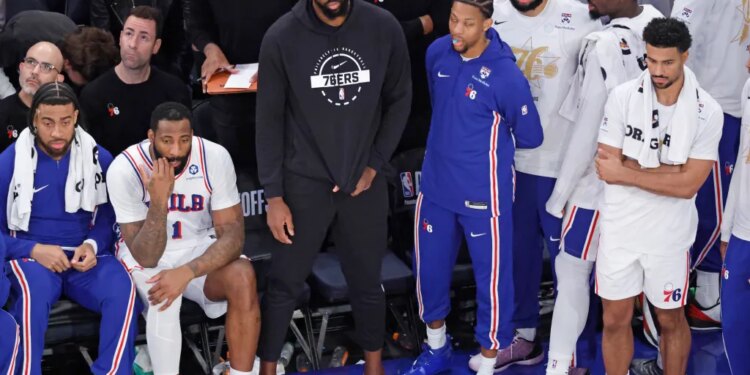 Philadelphia 76ers center Joel Embiid sits on the bench in plain clothes with teammates during a game.
